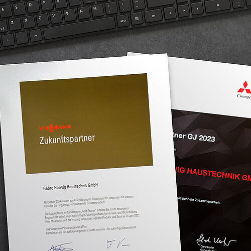 Eine goldene Urkunde mit der Auszeichnung "Zukunftspartner" von Viessmann und eine schwarze Urkunde für ausgezeichnete Zusammenarbeit der Firma Mitsubishi Electric liegen auf einem schwarzen Schreibtisch neben Mouse und Tastatur