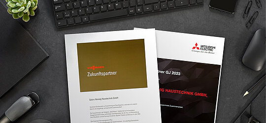 Eine goldene Urkunde mit der Auszeichnung "Zukunftspartner" von Viessmann und eine schwarze Urkunde für ausgezeichnete Zusammenarbeit der Firma Mitsubishi Electric liegen auf einem schwarzen Schreibtisch neben Mouse und Tastatur 