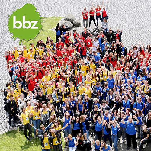 Eine große Gruppe an jungen Menschen in blau, gelben oder roten Tshirts, die die Arme in die Höhe strecken und Jubeln. Das Foto wurde von oben aufgenommen.