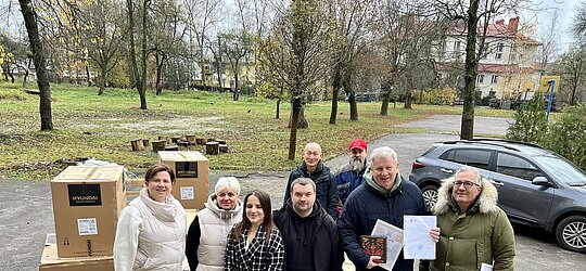 8 Personen stehen vor Kartons mit Generatoren, lächeln in die Kamera, im Hintergrund ein historisches Gebäude in der Stadt Lviv/Ukraine. lächeln 