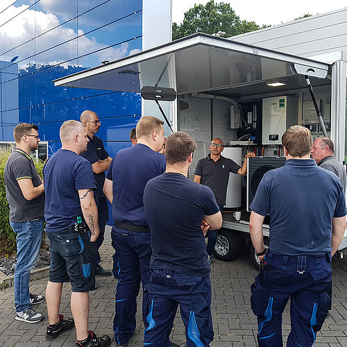 Zwei Vaillant Mitarbeiter präsentieren eine Wärmepumpe in einem Musteraufbau in einem dafür eingerichteten Anhänger. 8 Mitarbeiter von GEBRO HERWIG stehen im Halbkreis und hören interessiert zu.