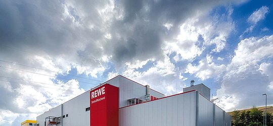 Eine Rückseite von einem Rewe-Geschäft, welches eine graue Fassade hat und rote Highlights sowie die Aufschrift Rewe.