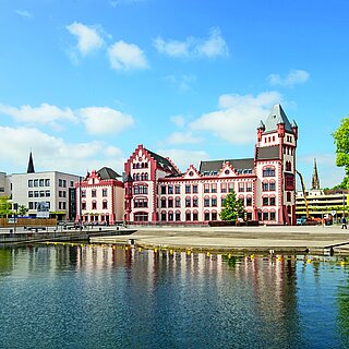 Ein helles Gebäude mit roten Umrandungen der Ecken und Fenster am Wasser.