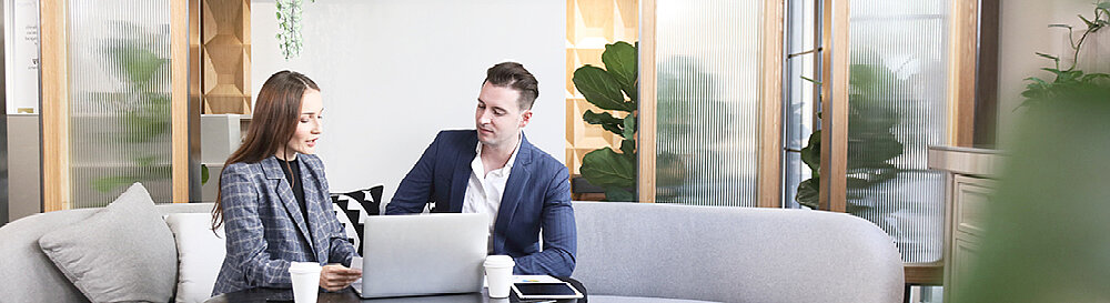 Ein Mann und eine Frau sitzen auf einem Sofa in einem Besprechungsraum, vor ihnen steht ein silberner Laptop und zwei Kaffeebecher To-Go. Die Blicke sind auf den Laptop gerichtet und die Personen haben einander zugewandt.